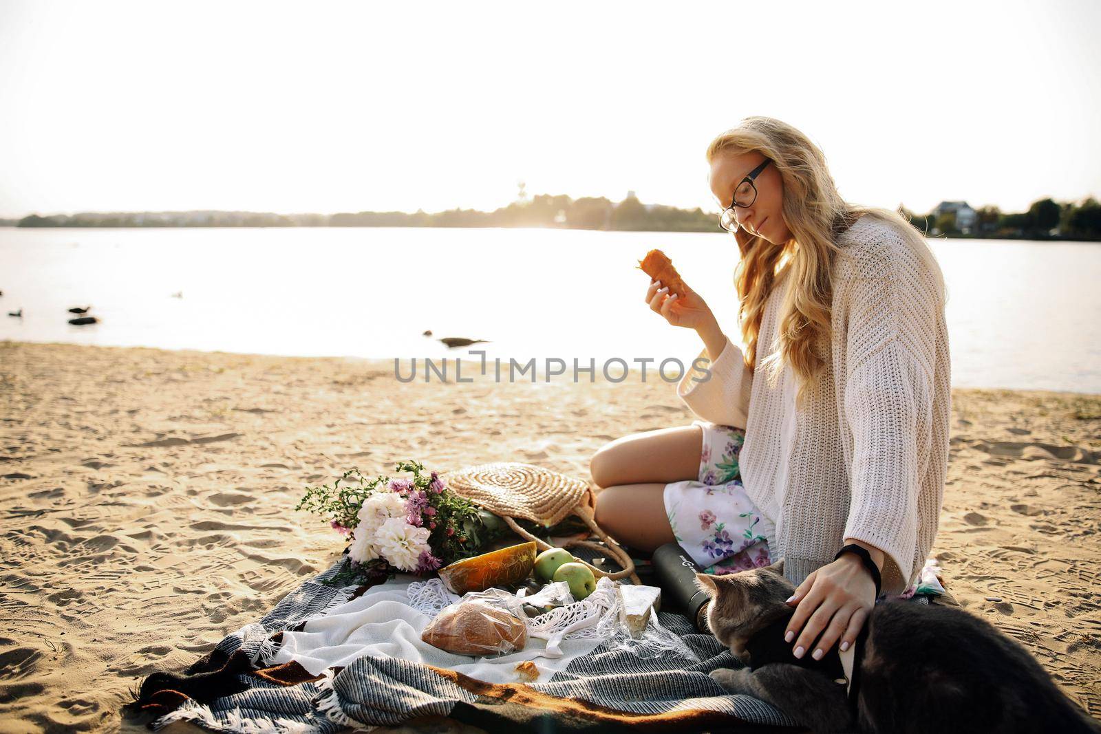 Young blonde woman lying on the beach in autumn in a white knitted sweater with a cat on a sunny day on a picnic
