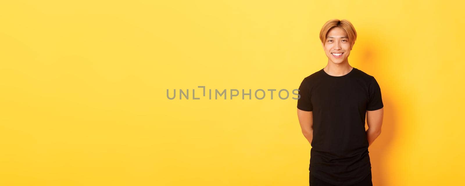 Portrait of friendly handsome asian guy with blond hair, smiling politely, holding hands behind back, standing yellow background.