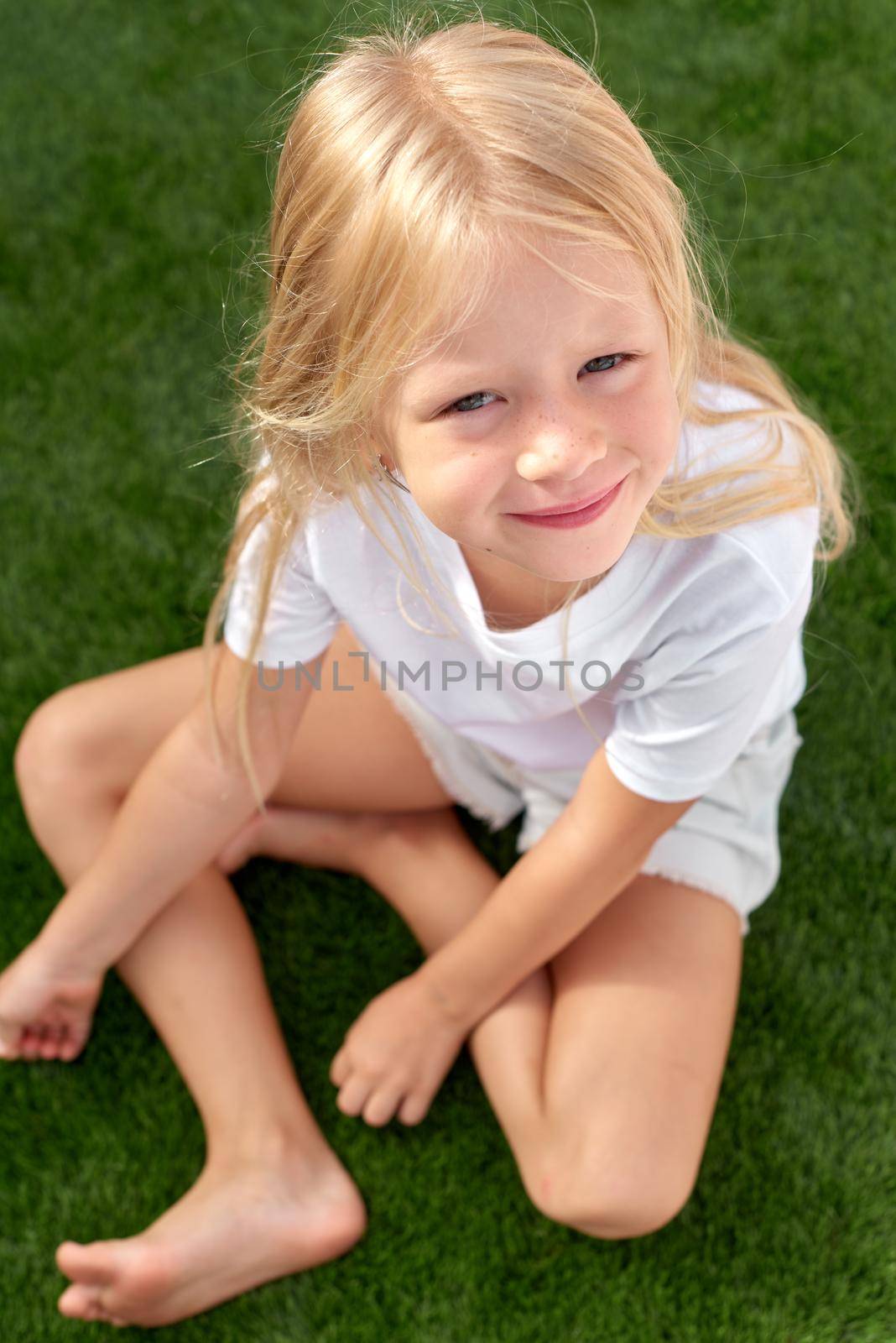 Mock up. Pretty beautiful blonde child girl sitting on green grass. Outdoors. Smiling preschool girl 5 - 6 years old in white t shirt looking at camera. Summer vacations. Leisure. People concept