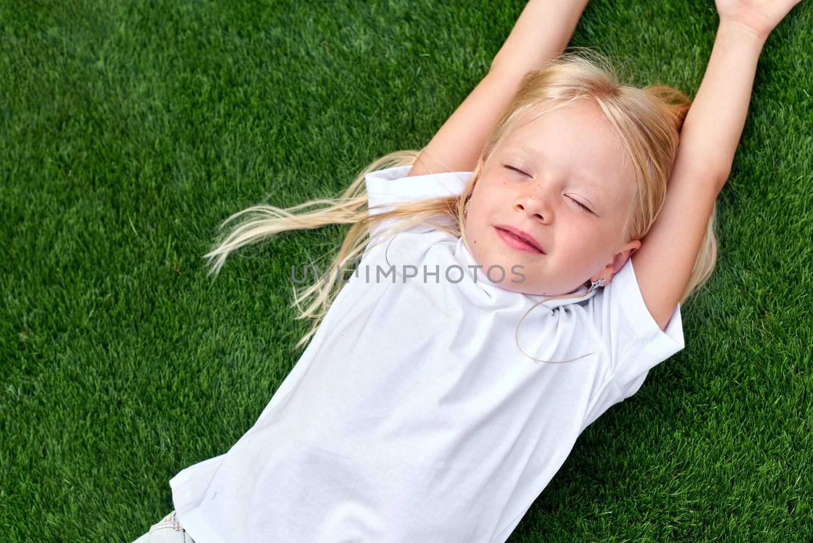Top view. Mock up for logo, text, design. Blonde child girl closed eyes lying stretching on green grass. Preschool girl 5-6 years old in white t shirt. Lifestyle Summer vacations Leisure. People