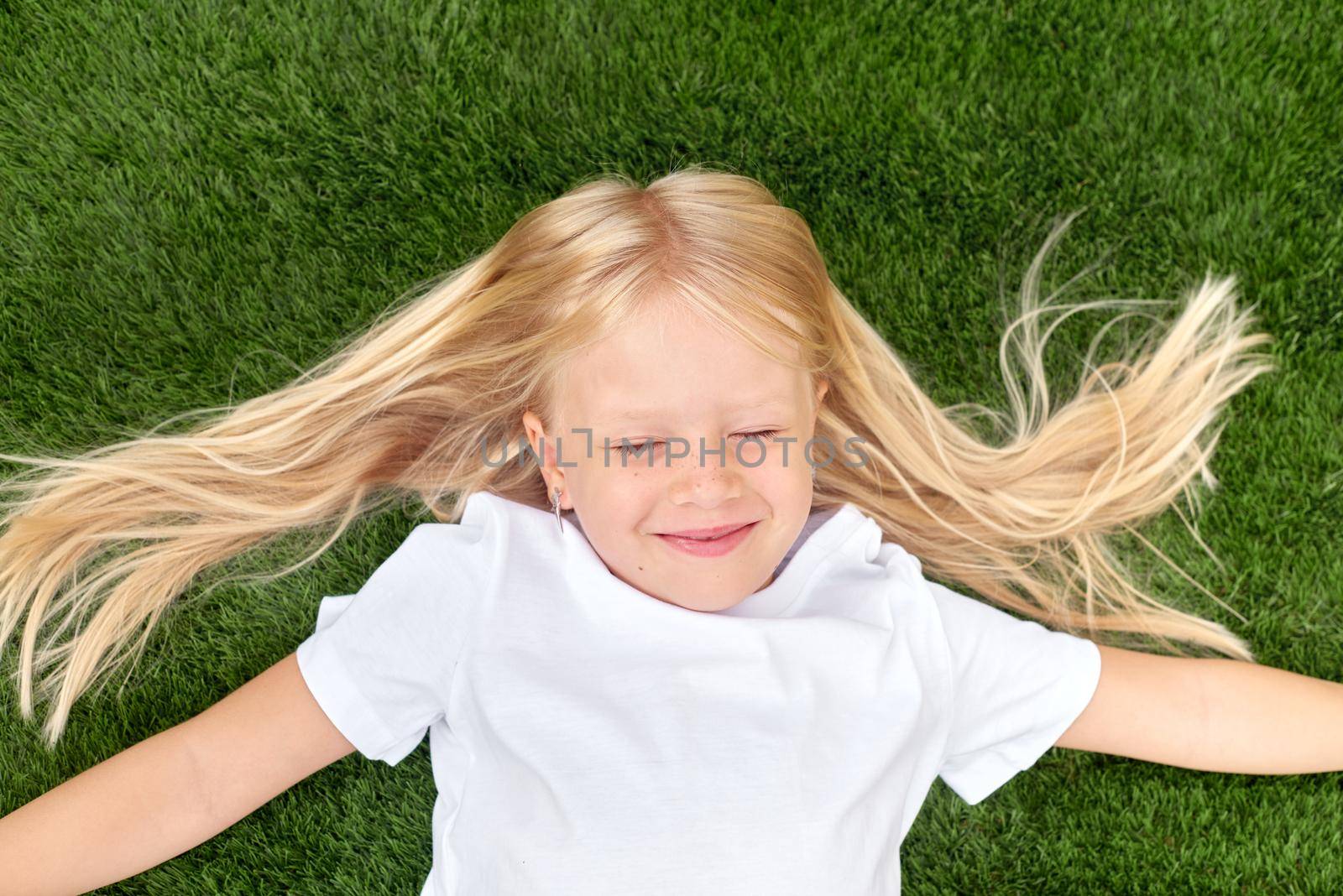 Top view. Mock up. Pretty beautiful blonde child girl lying and stretching on green grass. Preschool girl 5-6 years old in white t shirt. Lifestyle Summer vacations Leisure. People concept