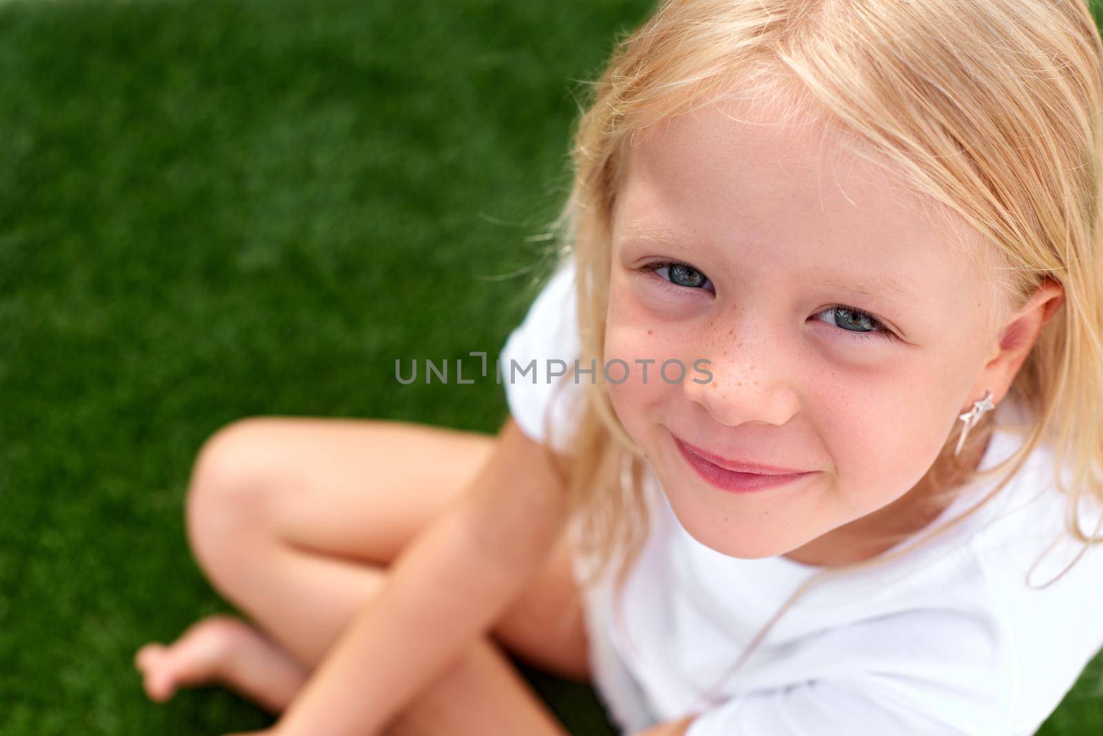Top view. Pretty beautiful blonde child girl sitting on green grass. Smiling preschool girl 5 - 6 years old in white t shirt looking at camera. Lifestyle. Beauty. Summer vacations. leisure. people