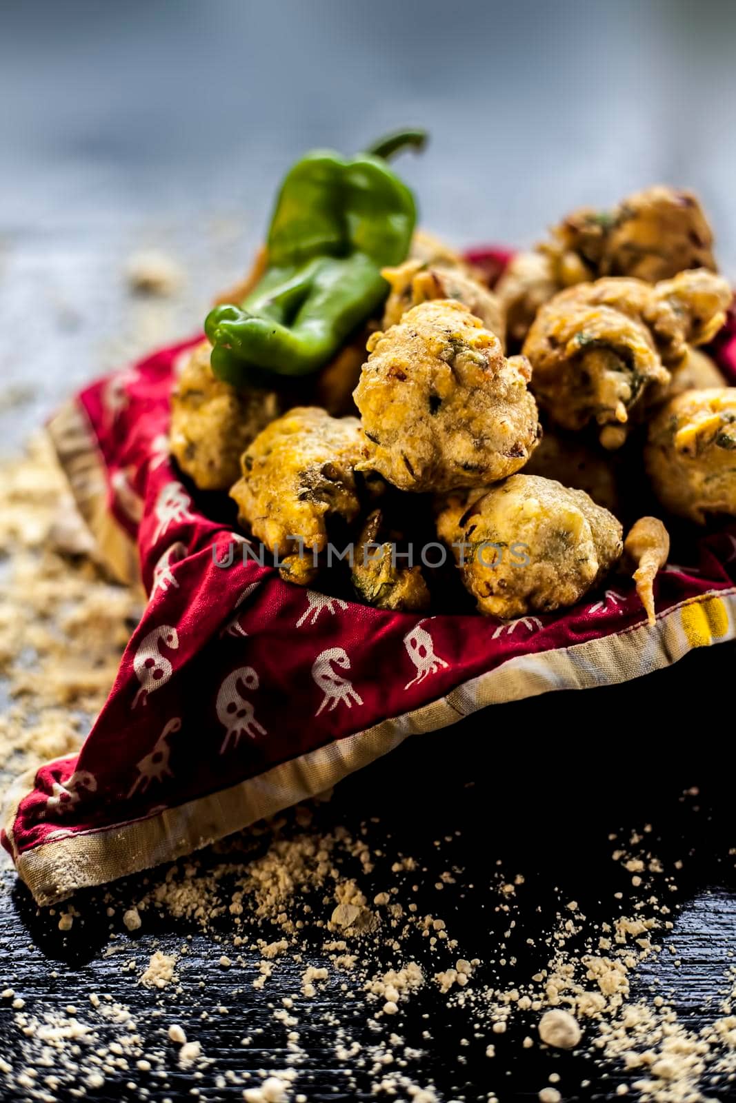 Close up shot of freshly fried methi pakora in a container on a black surface along with some spices and a chickpea flour spread on the surface. Methi pakora in a serving dish on the black surface.