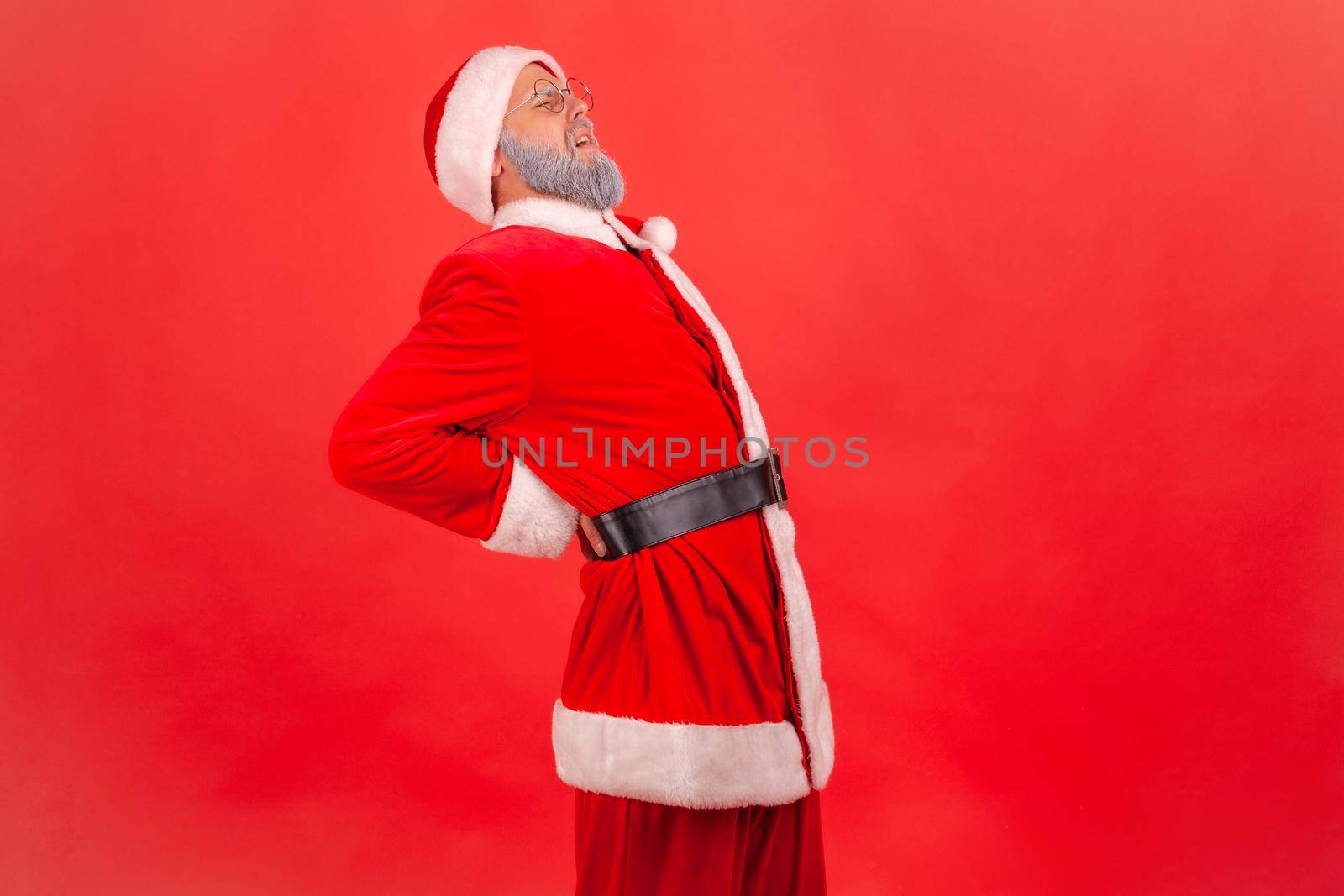 Sick elderly man with gray beard wearing santa claus costume feeling sharp pain in kidneys, back injury, pinched sciatic nerve, suffering. Indoor studio shot isolated on red background.