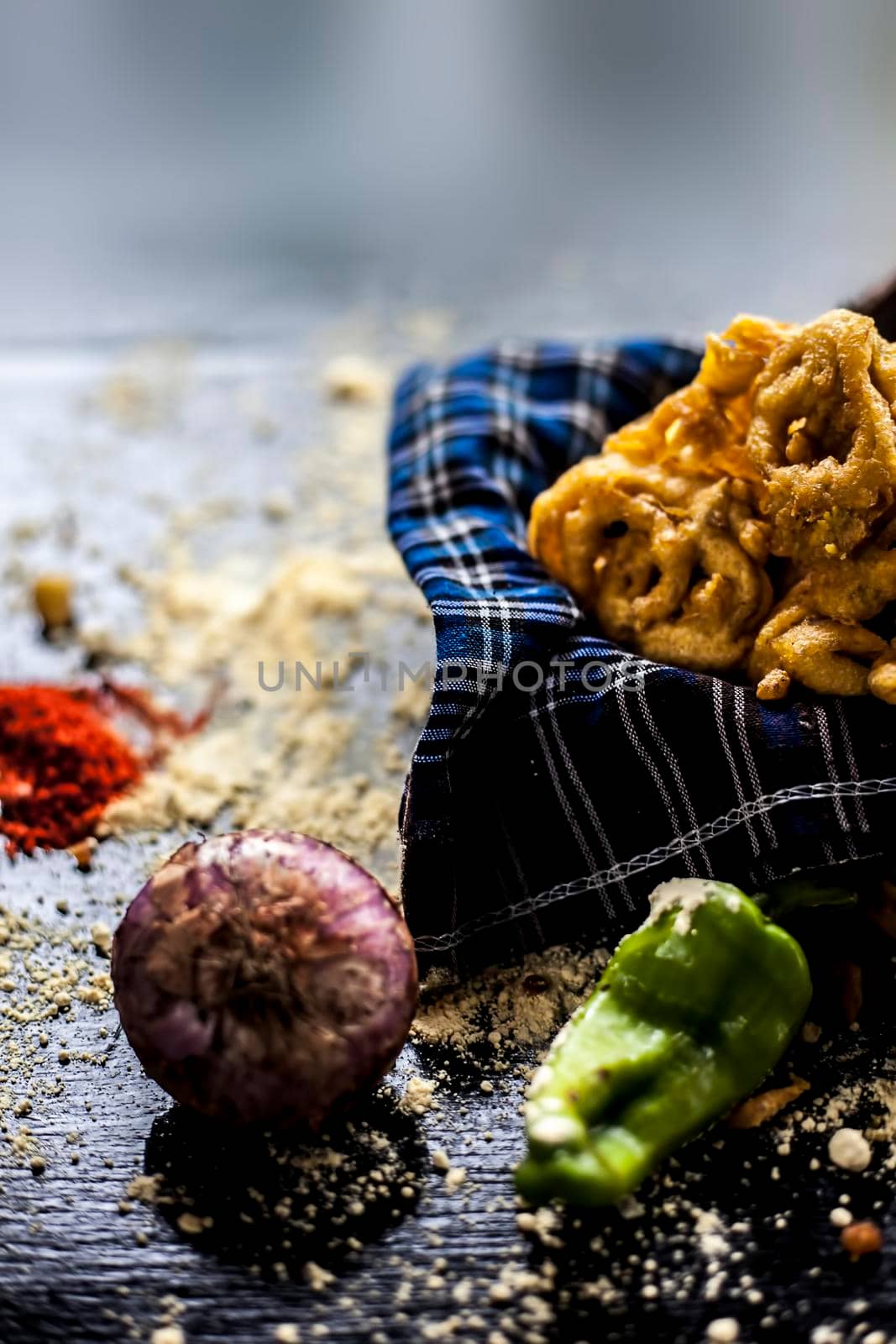 Famous Kanda bhaji or Kanda bhajiya or Kanda pakora in a container on a black surface along with chickpea flour, spices, onion, and all other ingredients for making it. Vertical shot.