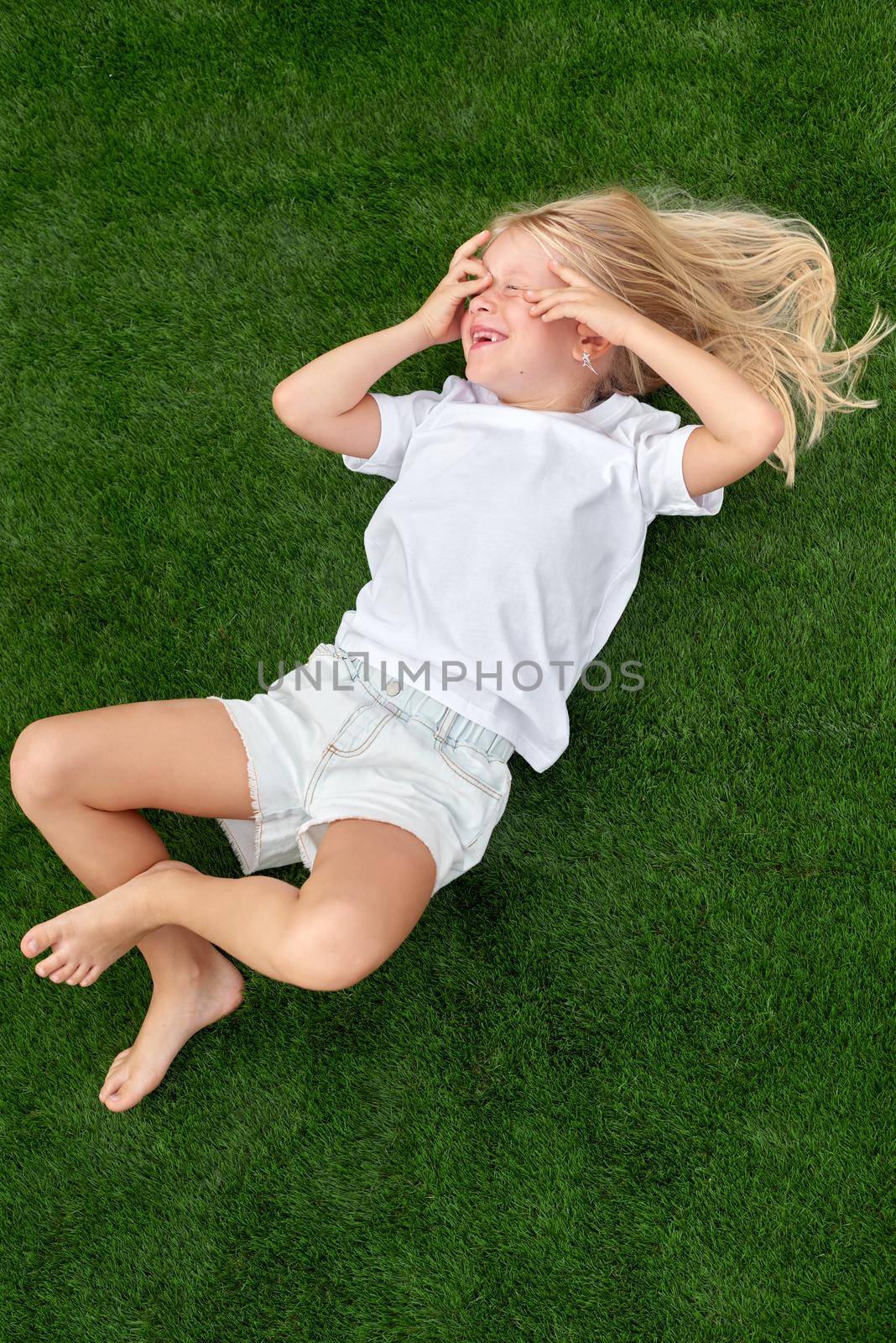 Top view. Mock up. Cute beautiful blonde child girl lying and stretching on green grass. Smiling preschool girl 5-6 years old in white t shirt. Lifestyle Summer vacations Leisure. People concept