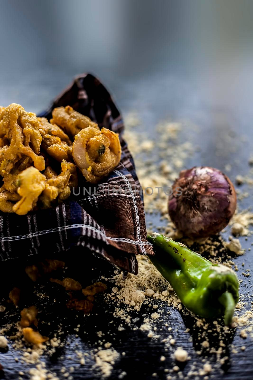 Famous Kanda bhaji or Kanda bhajiya or Kanda pakora in a container on a black surface along with chickpea flour, spices, onion, and all other ingredients for making it. Vertical shot.