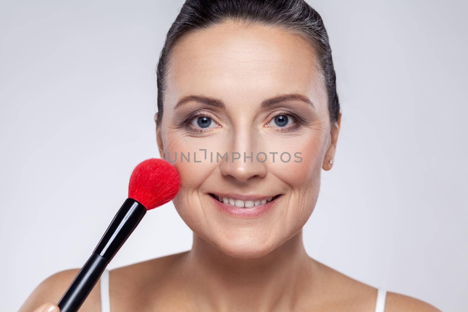 Closeup portrait of beautiful smiling middle aged woman with perfect skin applying makeup with a red brush, preparing for an important event. Indoor studio shot isolated on gray background.