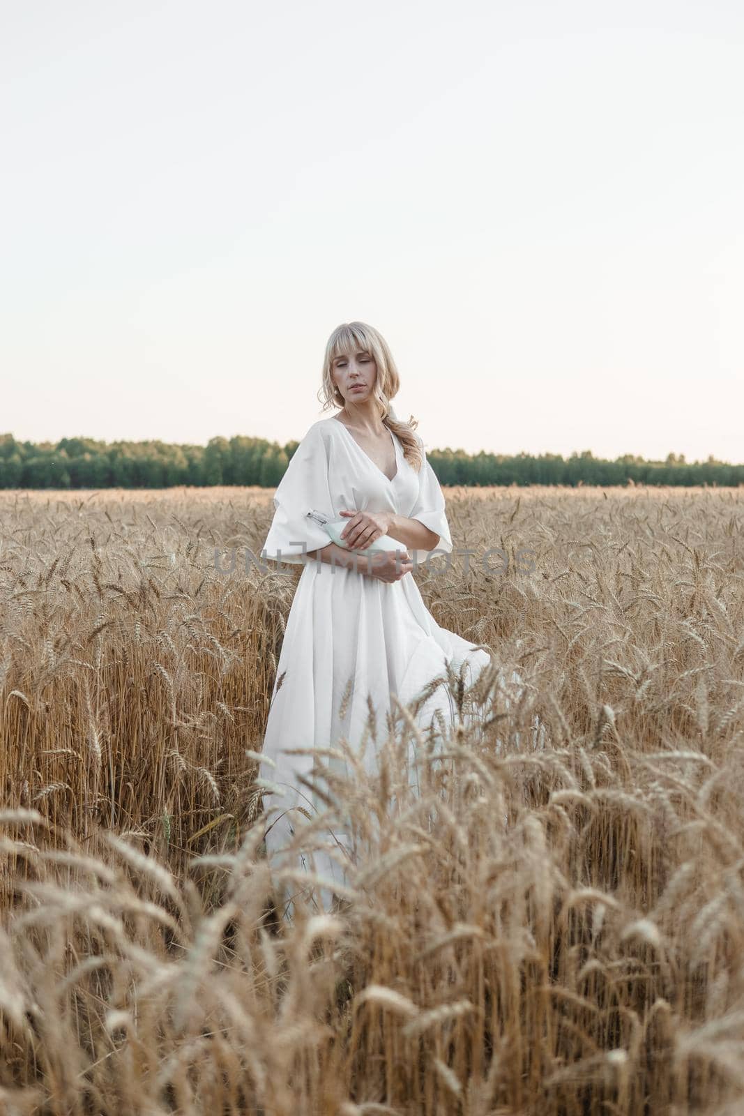 A blonde woman in a long white dress walks in a wheat field. The concept of a wedding and walking in nature.