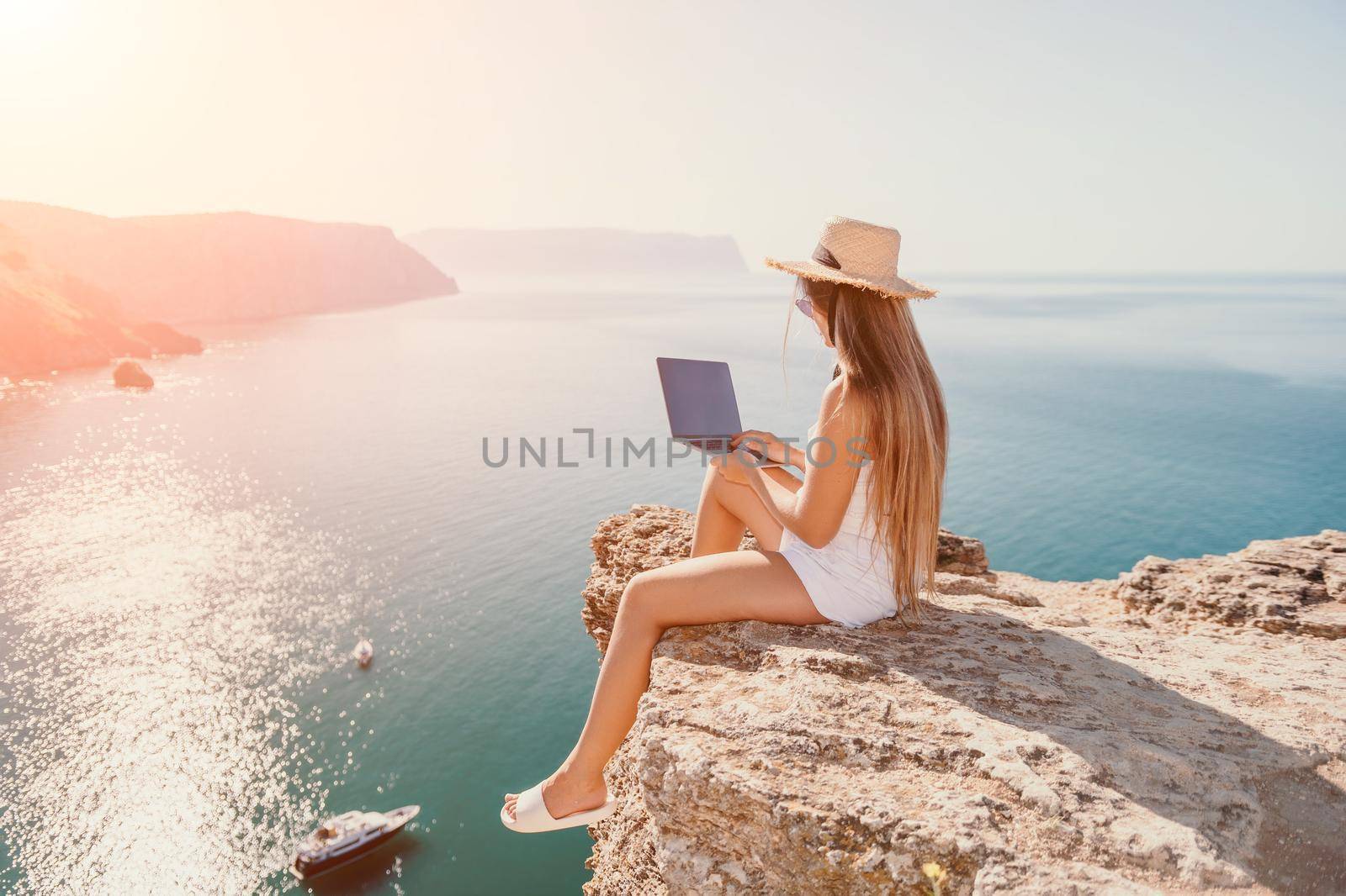 Successful business woman in yellow hat working on laptop by the sea. Pretty lady typing on computer at summer day outdoors. Freelance, travel and holidays concept.