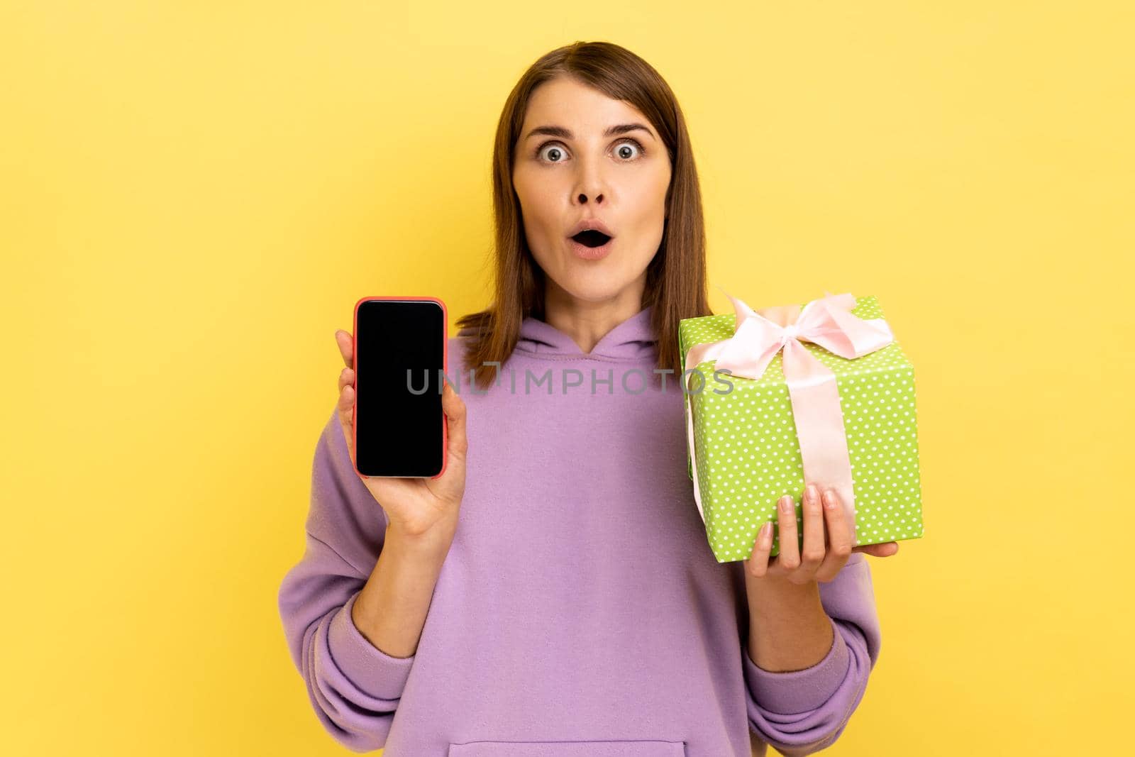 Present, bonus for mobile user. Portrait of amazed surprised woman holding gift box and cell phone with mock up, blank display, wearing hoodie. Indoor studio shot isolated on yellow background.