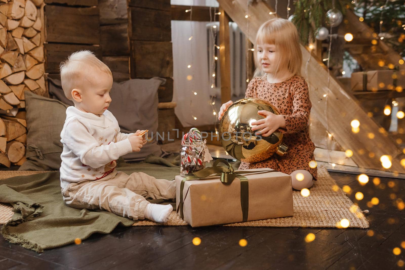 Little brother and sister play on Christmas eve in a beautiful house decorated for the New Year holidays. Children are playing with a Christmas gift. Scandinavian-style interior with live fir trees and a wooden staircase.