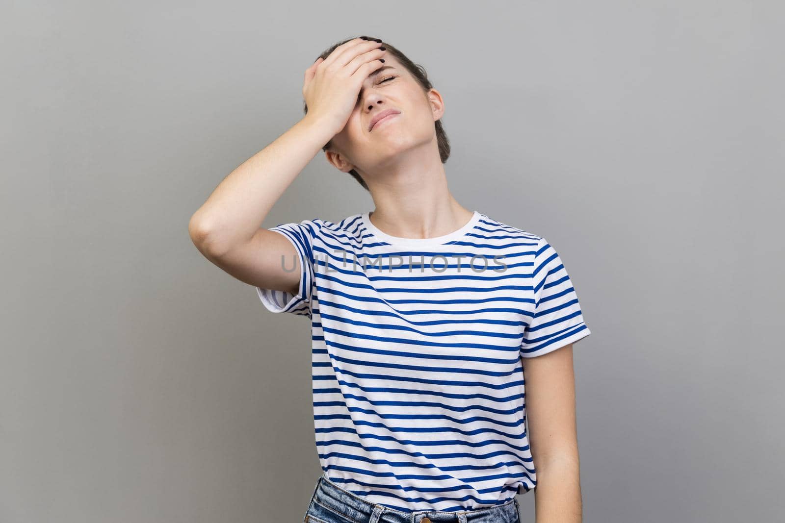 Portrait of upset frustrated woman wearing striped T-shirt holding forehead with hand, making facepalm gesture, forget about date, big failure. Indoor studio shot isolated on gray background.