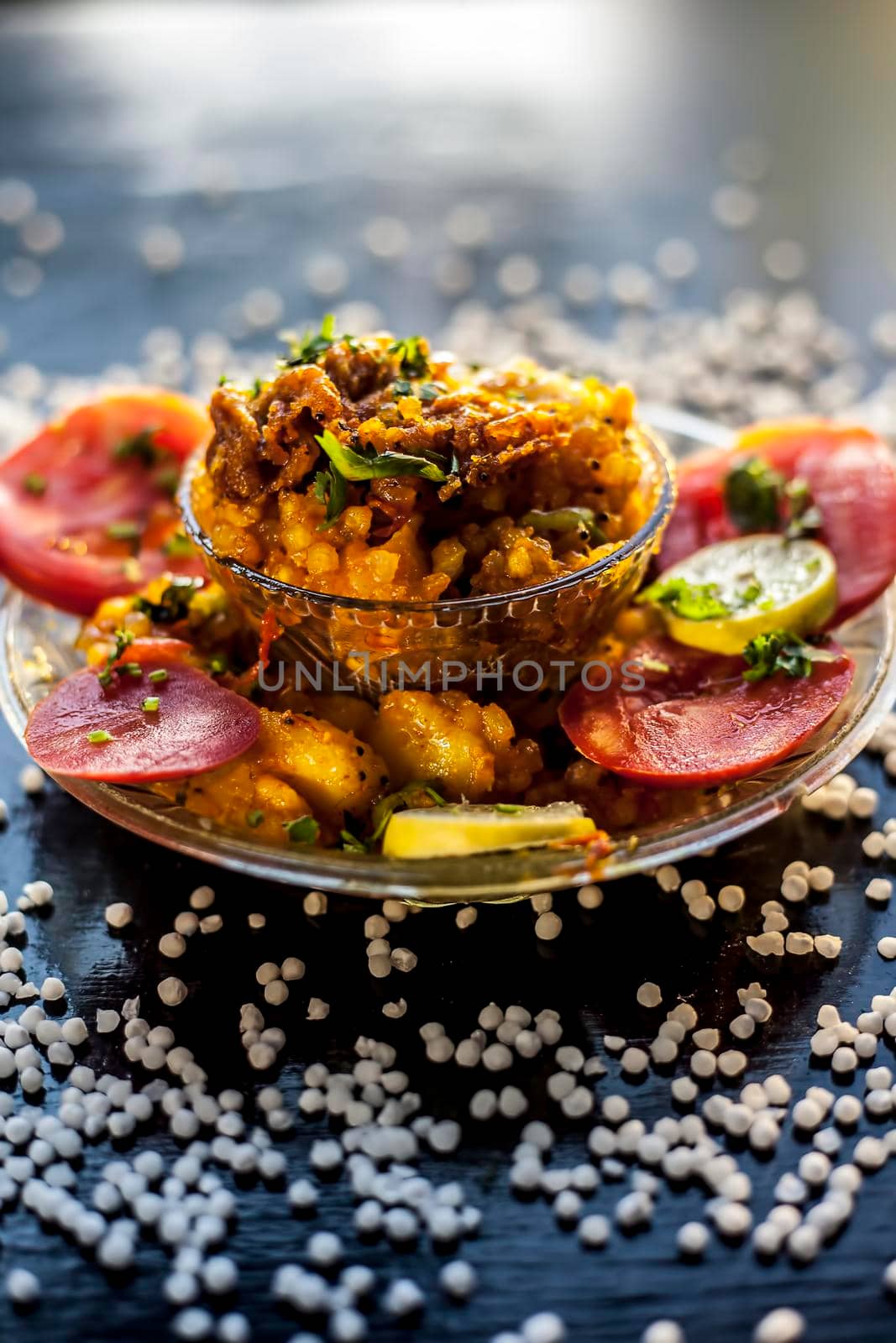 Close up shot of spicy tasty sabudana khichdi or sago ball khichdi along with some sliced tomatoes, some cut lemons in a glass plate, and raw sago balls.