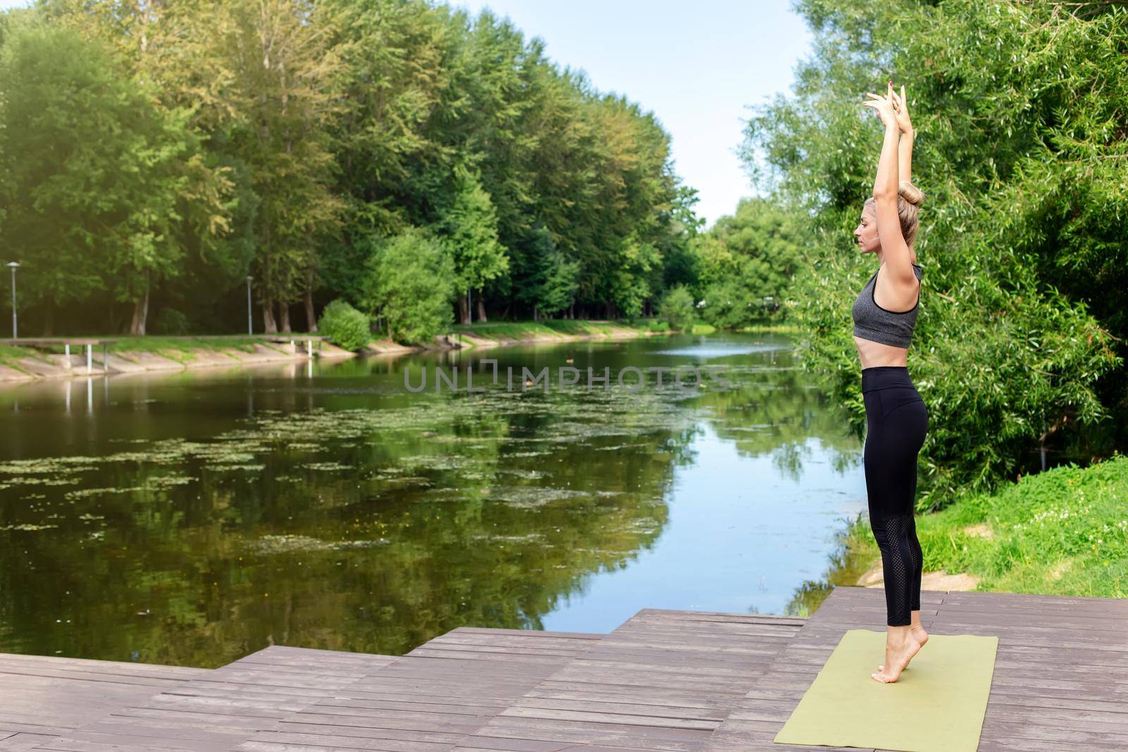 A slender woman stands on a green rug, on a wooden platform by a pond in the park, in summer, with her hands up, doing yoga. Copy space