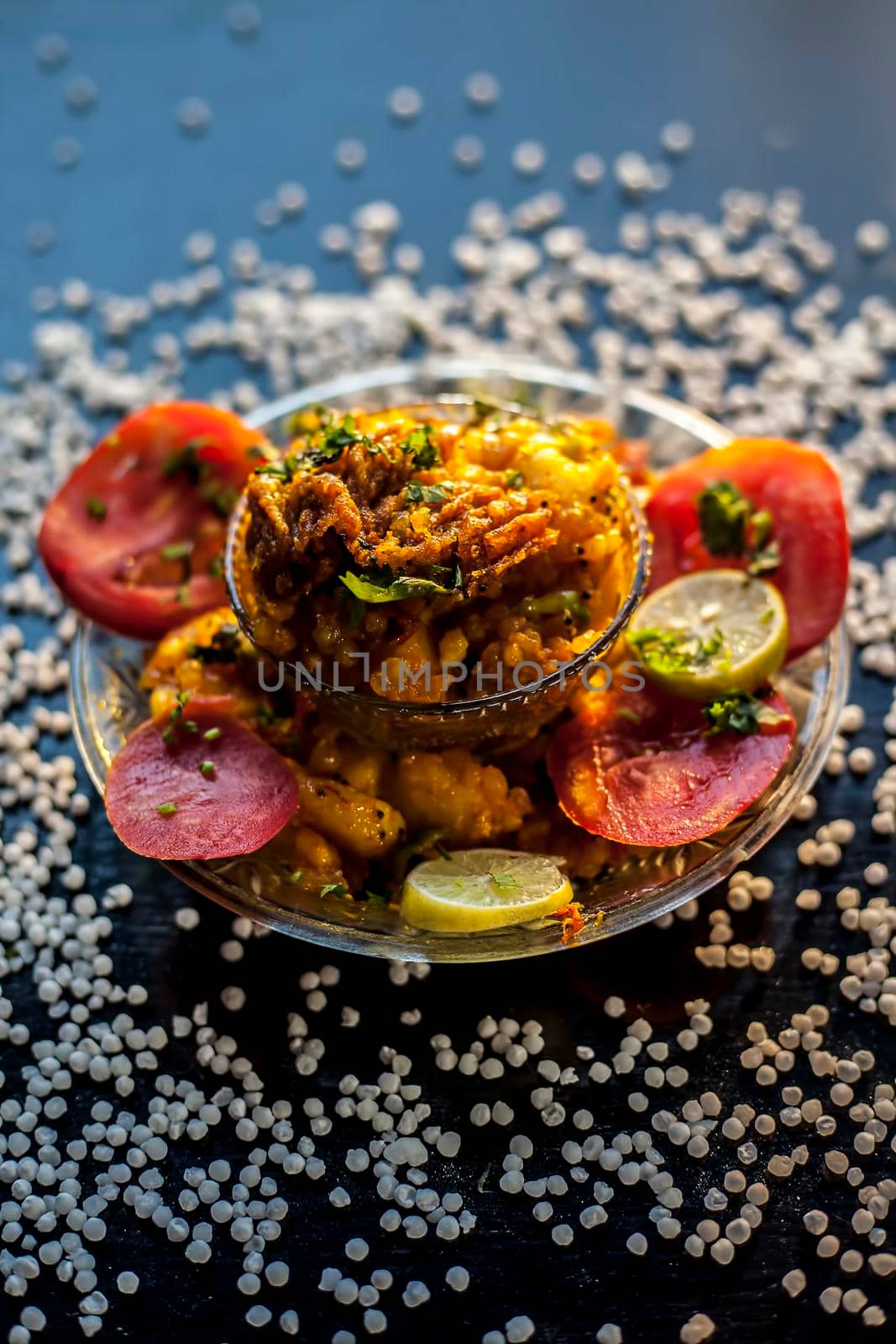 Close up shot of spicy tasty sabudana khichdi or sago ball khichdi along with some sliced tomatoes, some cut lemons in a glass plate, and raw sago balls.