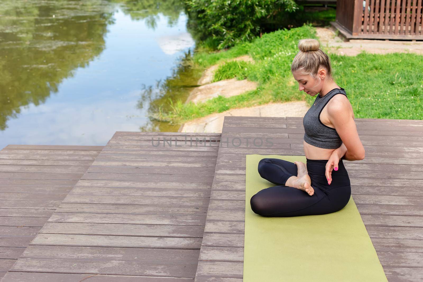 A slender woman in a gray spotrswear, sitting on a wooden platform by a pond in the park in summer and performing yoga. Copy space