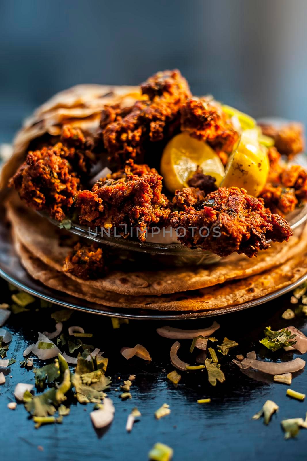 Shot of dry meatballs on top of the tortilla. Shot of kebab or kabab on roti along with some green chilies, lemon, and onion rings on a black surface.