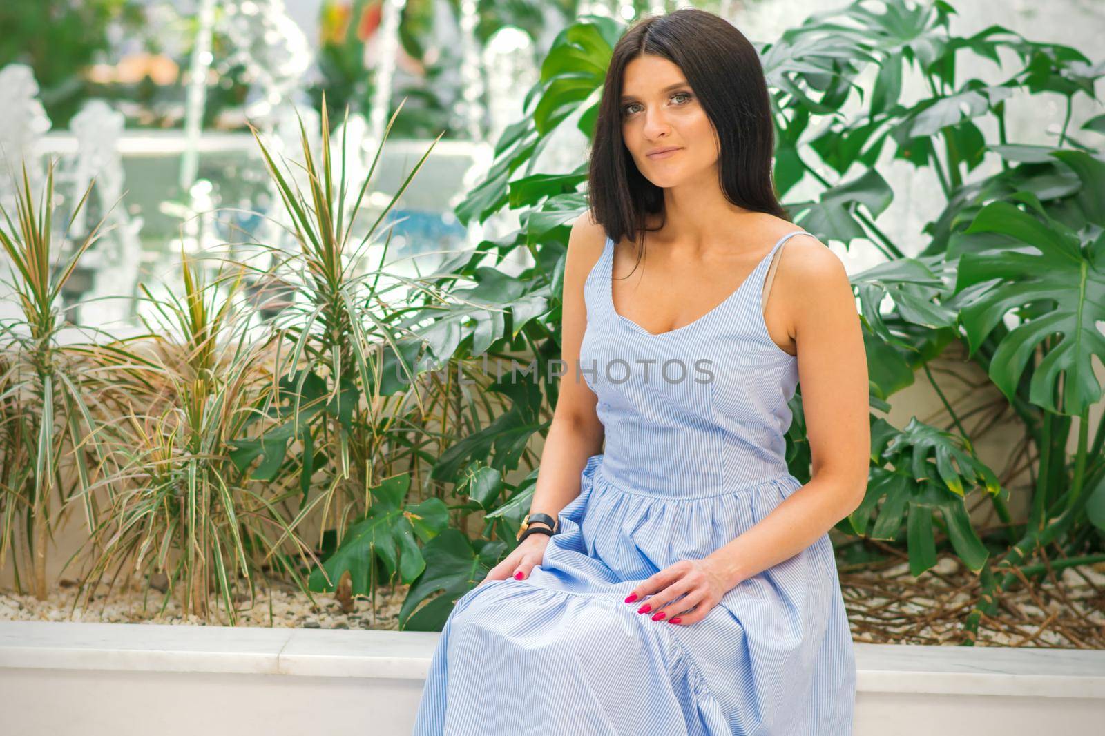 An outdoor portrait of an attractive brunette woman wearing a blue long dress is posing by the fountain at the park