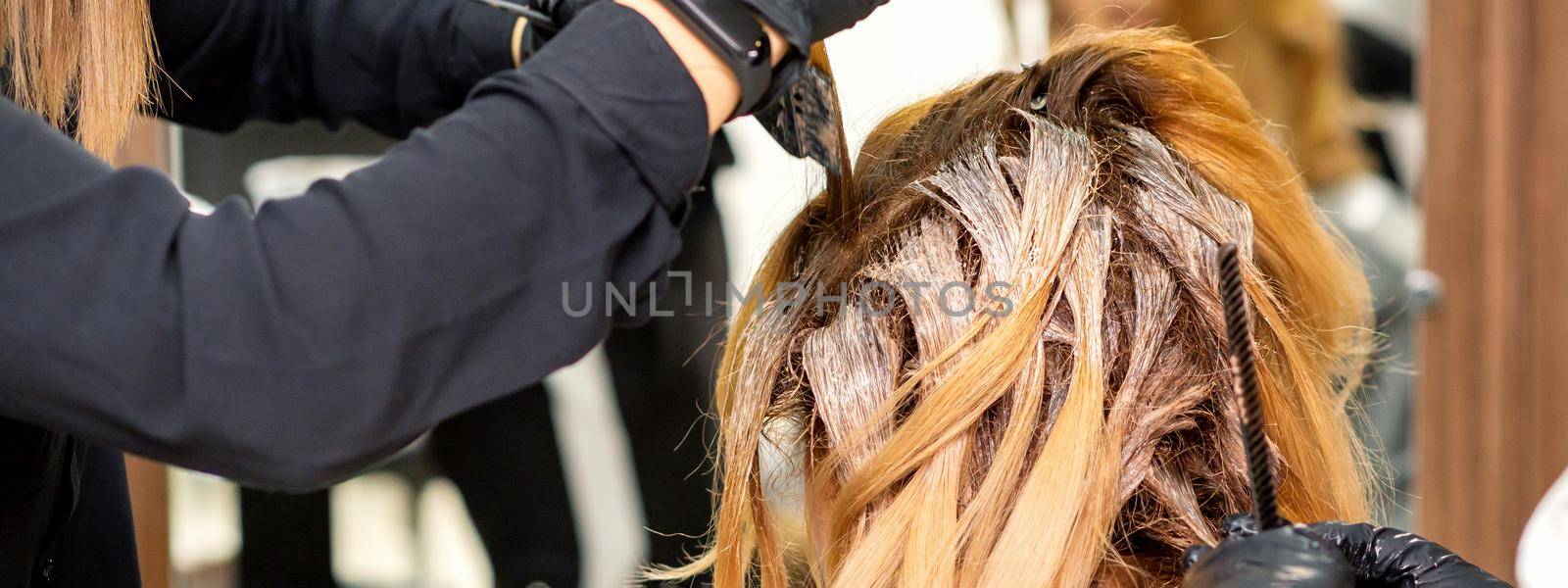 Back view of a female hairdresser in gloves coloring hair of redhead young woman with light or white dye in a beauty salon