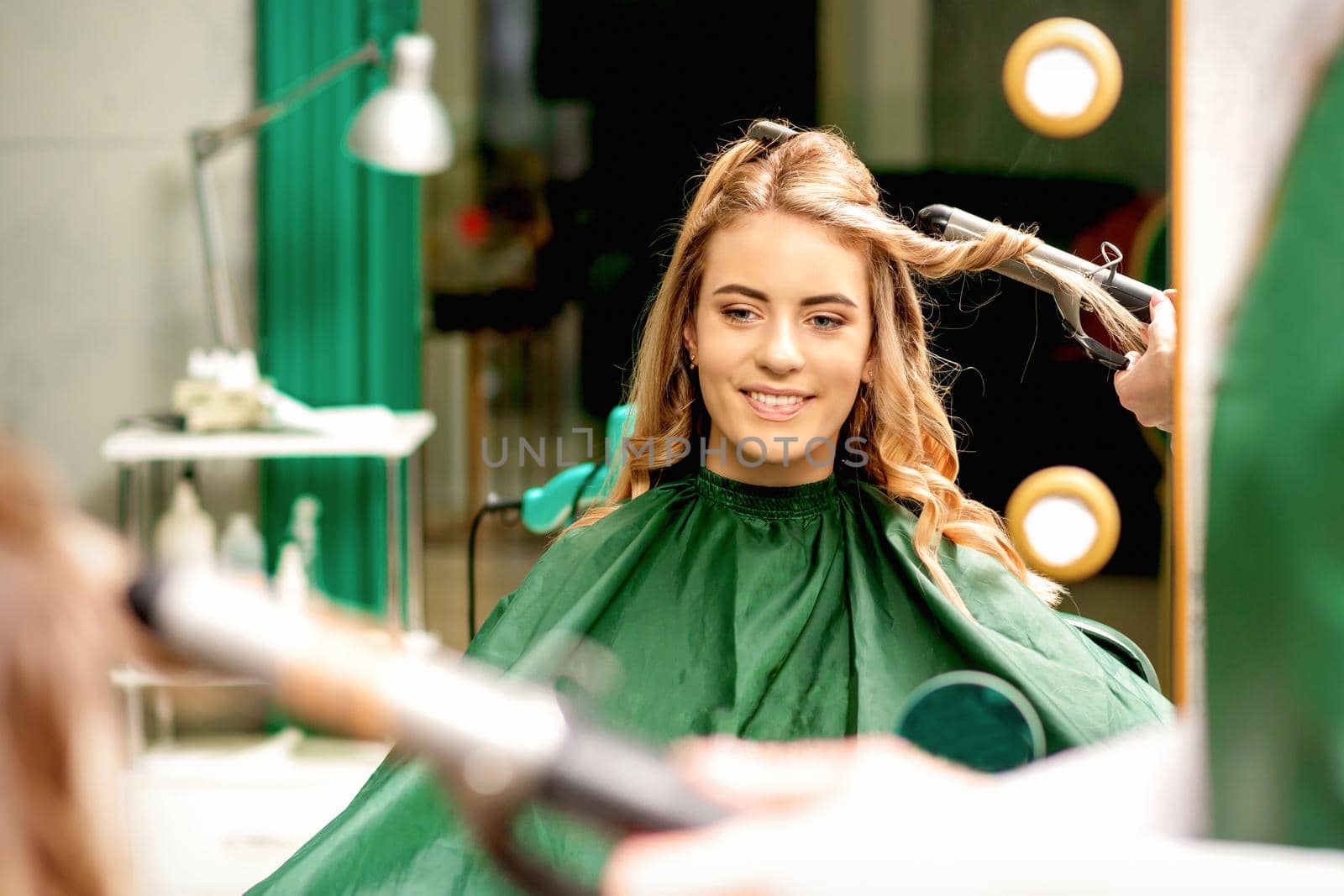 Hairdresser using curling tongs curls long brown hair on the young caucasian girl in a beauty salon