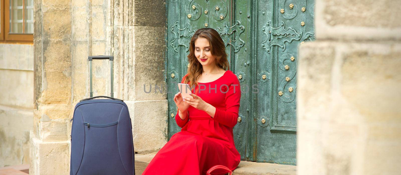 Beautiful young caucasian woman sitting on the stairs by the door with travel suitcase and smartphone wearing long red dress