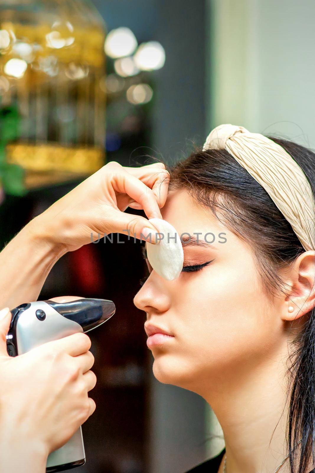 Side view of a make-up artist using aerograph making an airbrush makeup foundation on a female face in a beauty salon