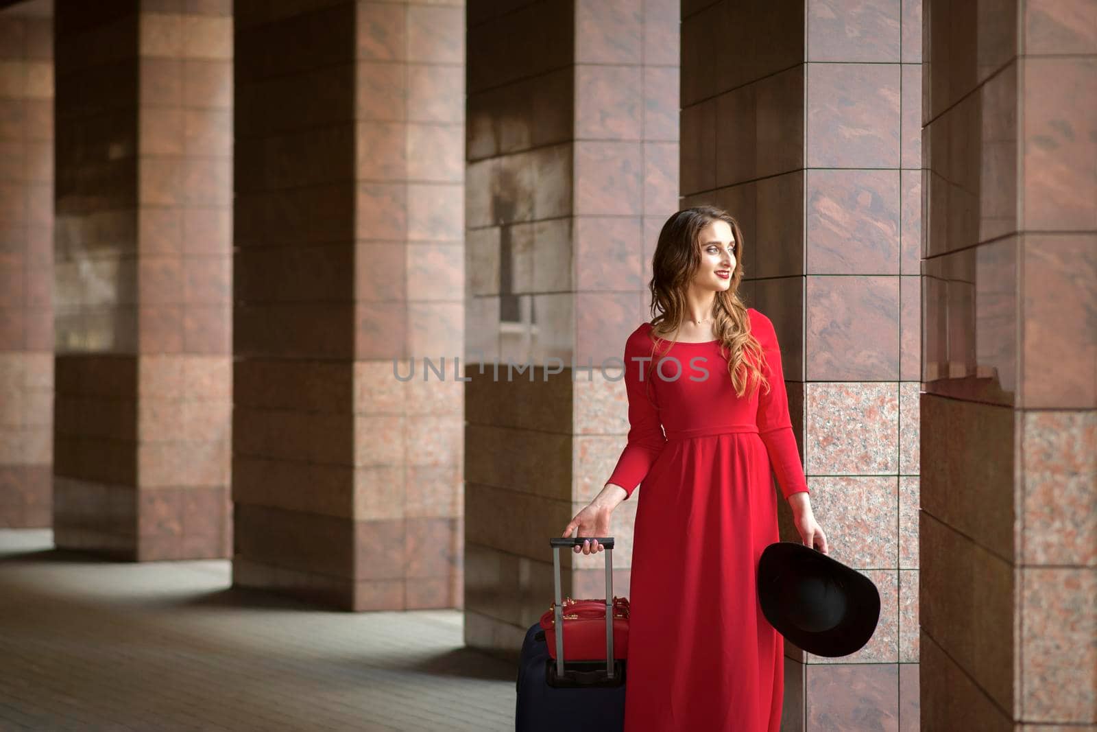 Beautiful young caucasian woman wearing red long dress with a suitcase standing in columns