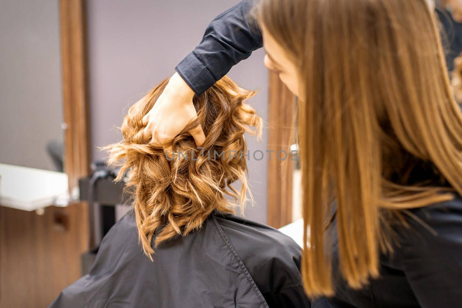 Female hairdresser checks brown curly hairstyle of a young caucasian woman in beauty salon