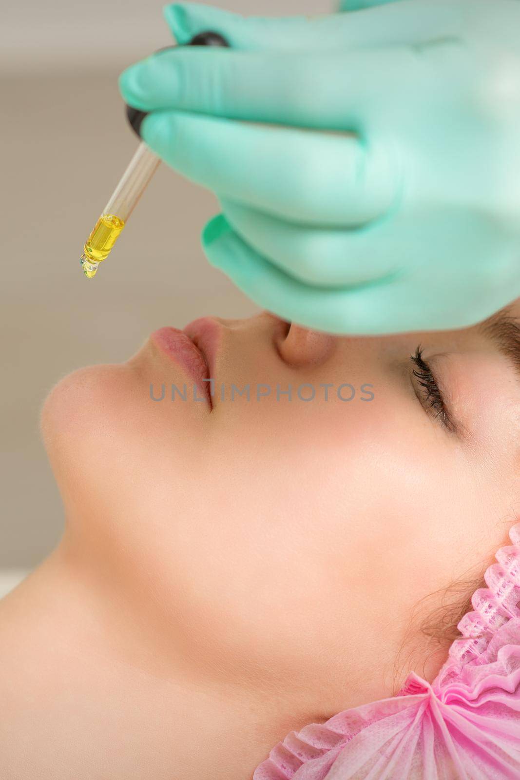 Close up of beautician holding pipette and applying oil serum drop on the face of a young woman in spa salon