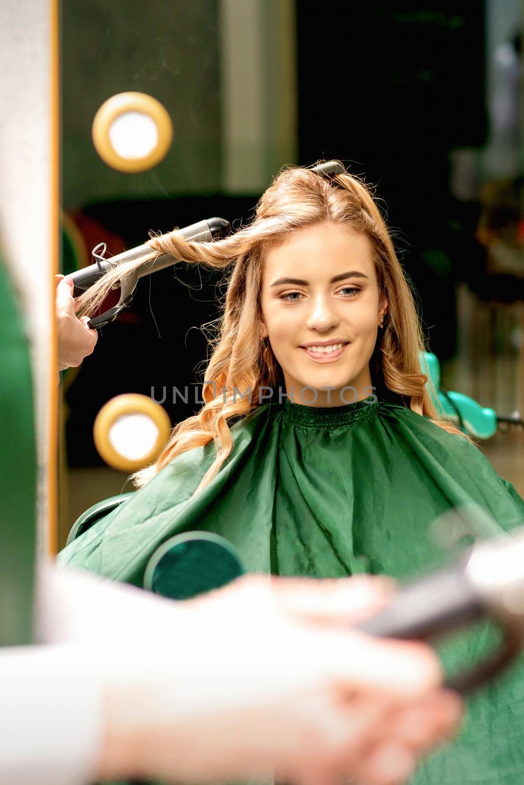 Hairdresser using curling tongs curls long brown hair on the young caucasian girl in a beauty salon