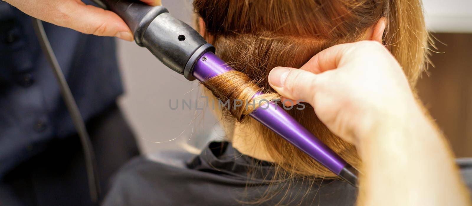 Close up of hairstylist's hands using a curling iron for hair curls in a beauty salon. by okskukuruza