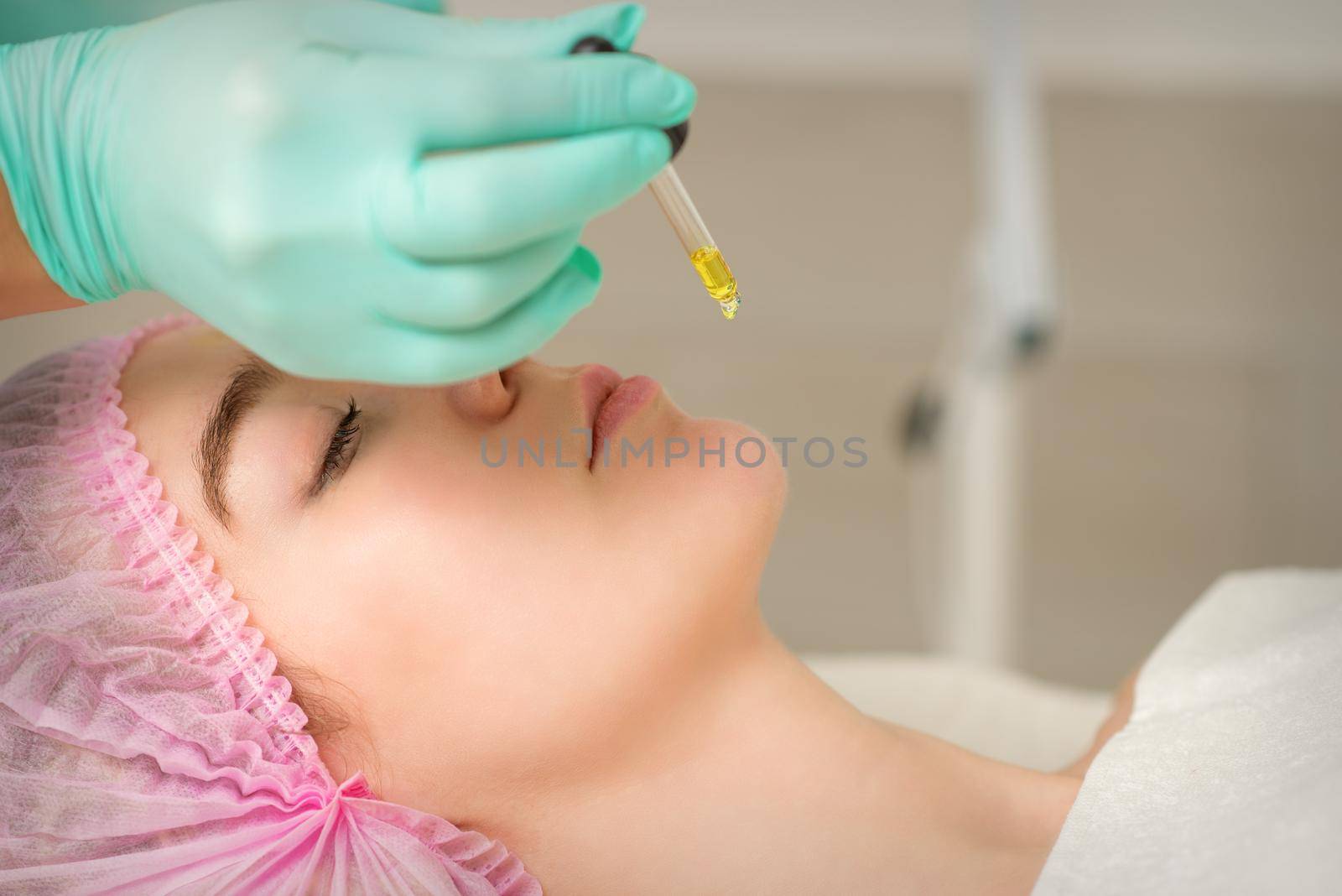 Close up of beautician holding pipette and applying oil serum drop on the face of a young woman in spa salon