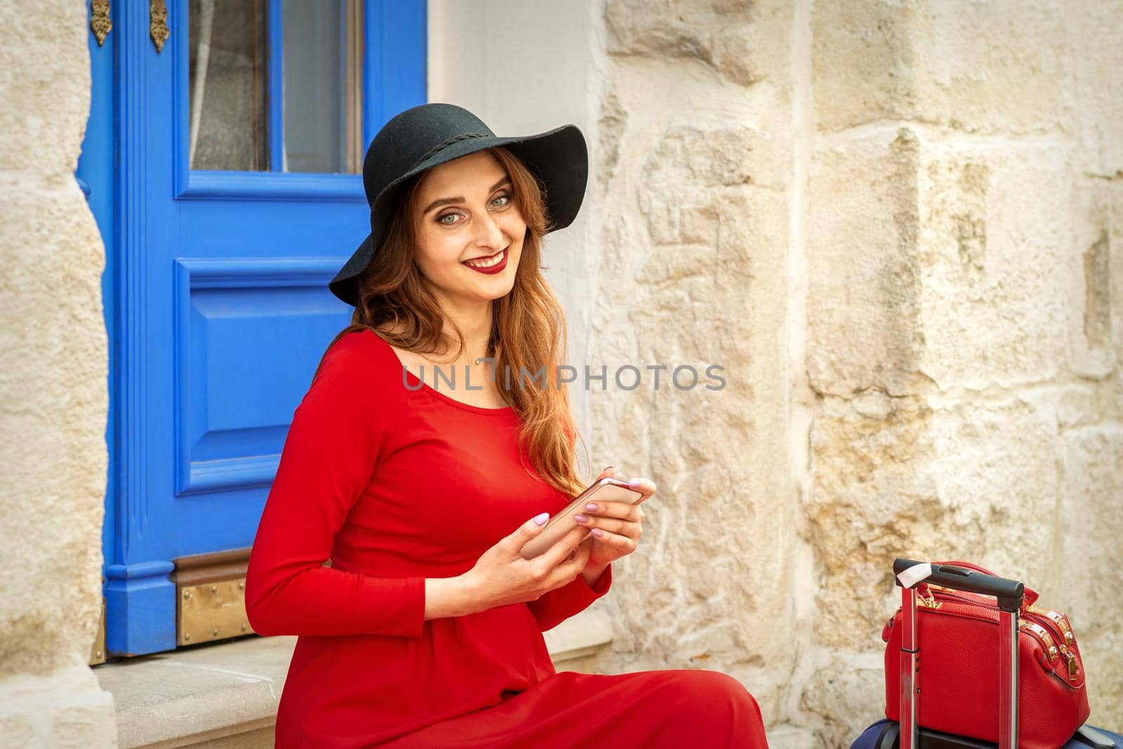 Portrait of tourist young caucasian woman in a red long dress and black hat with suitcase sitting on the stairs outdoors