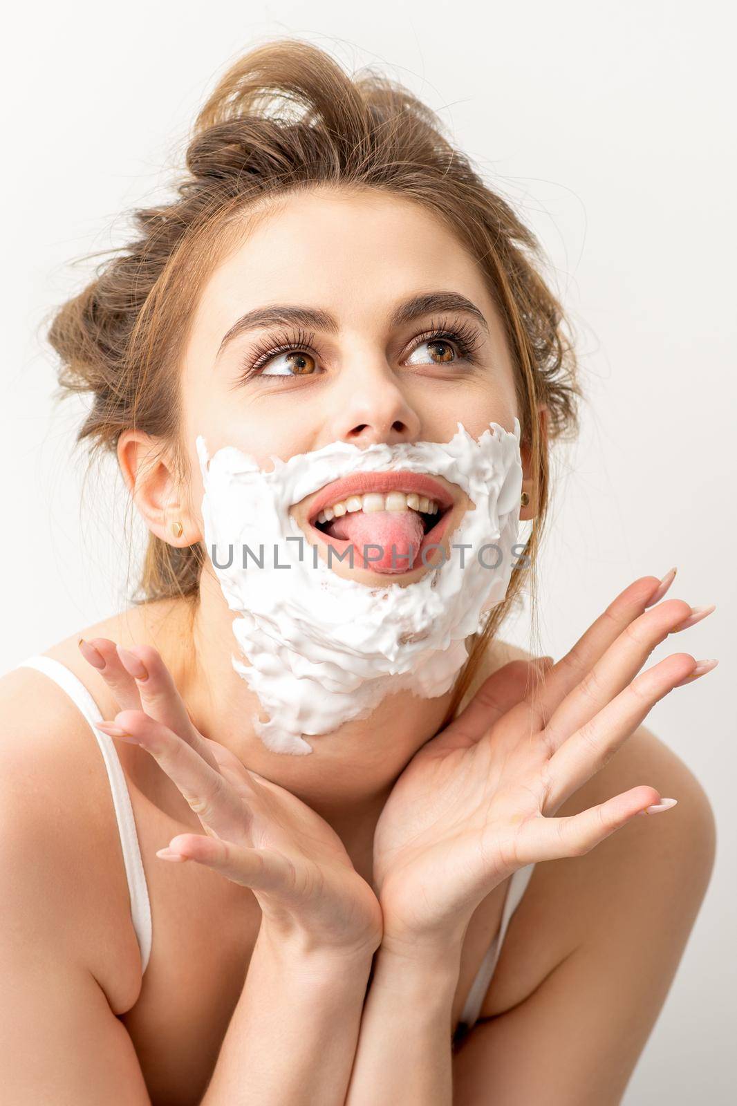 Portrait of a beautiful young smiling caucasian woman posing with shaving foam on the face stuck out her tongue on white background