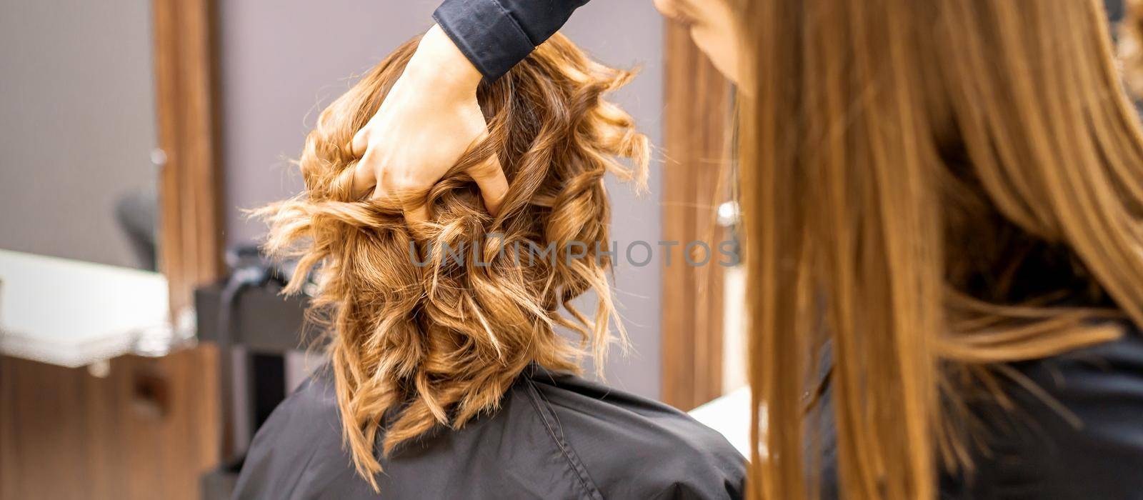 Female hairdresser checks brown curly hairstyle of a young caucasian woman in beauty salon. by okskukuruza