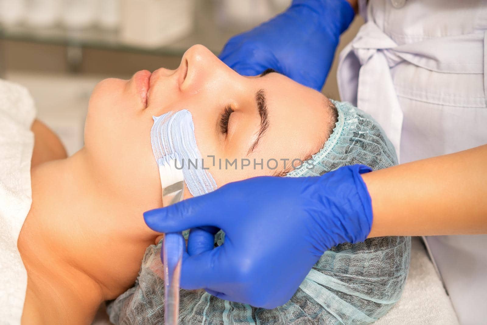 Beautician covering woman facial skin with moisturizing cleansing mask during skin care procedure in a spa beauty salon