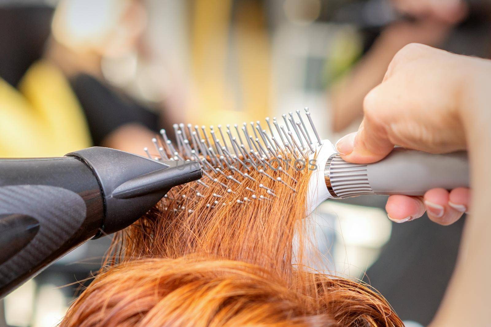 Closeup of master's hand with blow-drying and hairbrush blowing female red hair in a salon