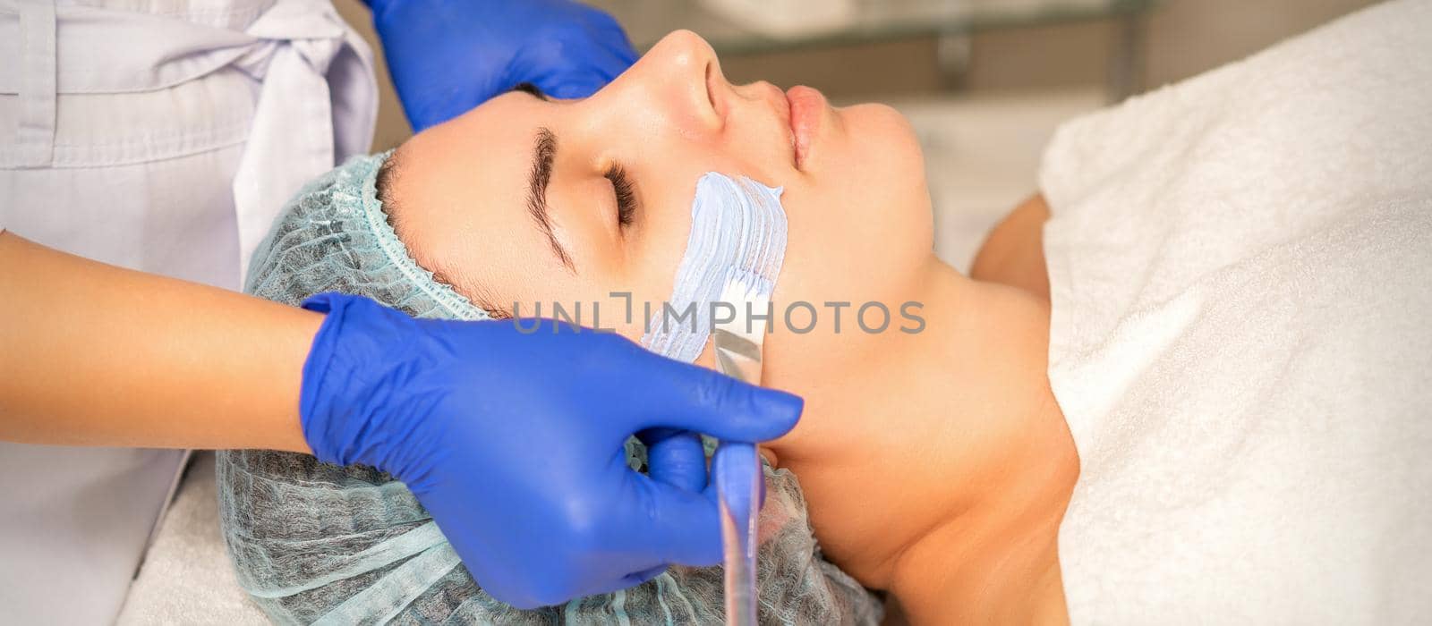 Beautician covering woman facial skin with moisturizing cleansing mask during skin care procedure in a spa beauty salon