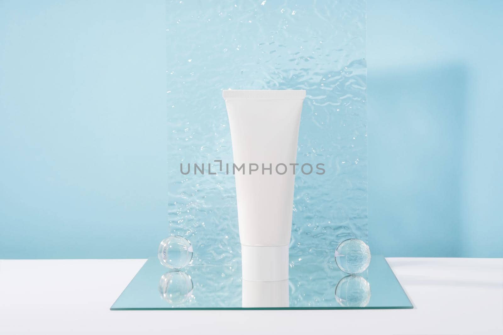 Unbranded cosmetic cream white plastic tube mockup on blue background with stylish props, mirror and glass balls, acrylic plate. Body and health care beauty product packaging by photolime