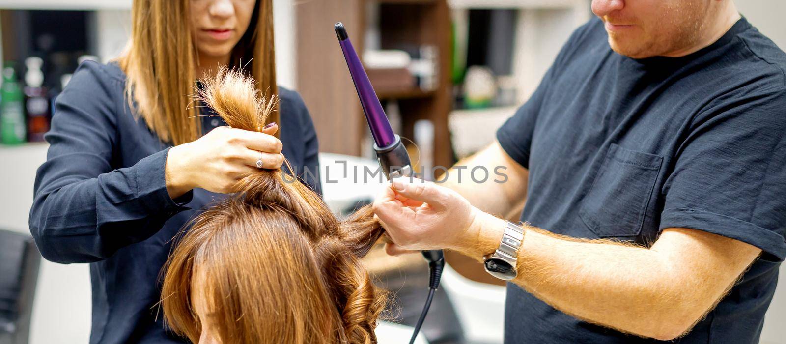 Two hairstylists using a curling iron on customers long brown hair in a beauty salon. by okskukuruza