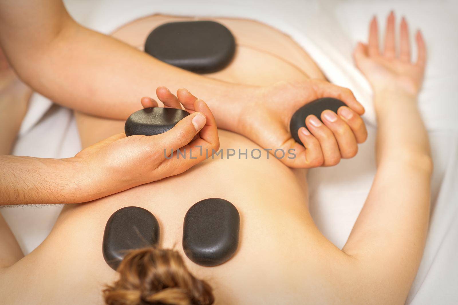 Hot stone massage on the female back with hands of masseur holding black massage stones in spa salon