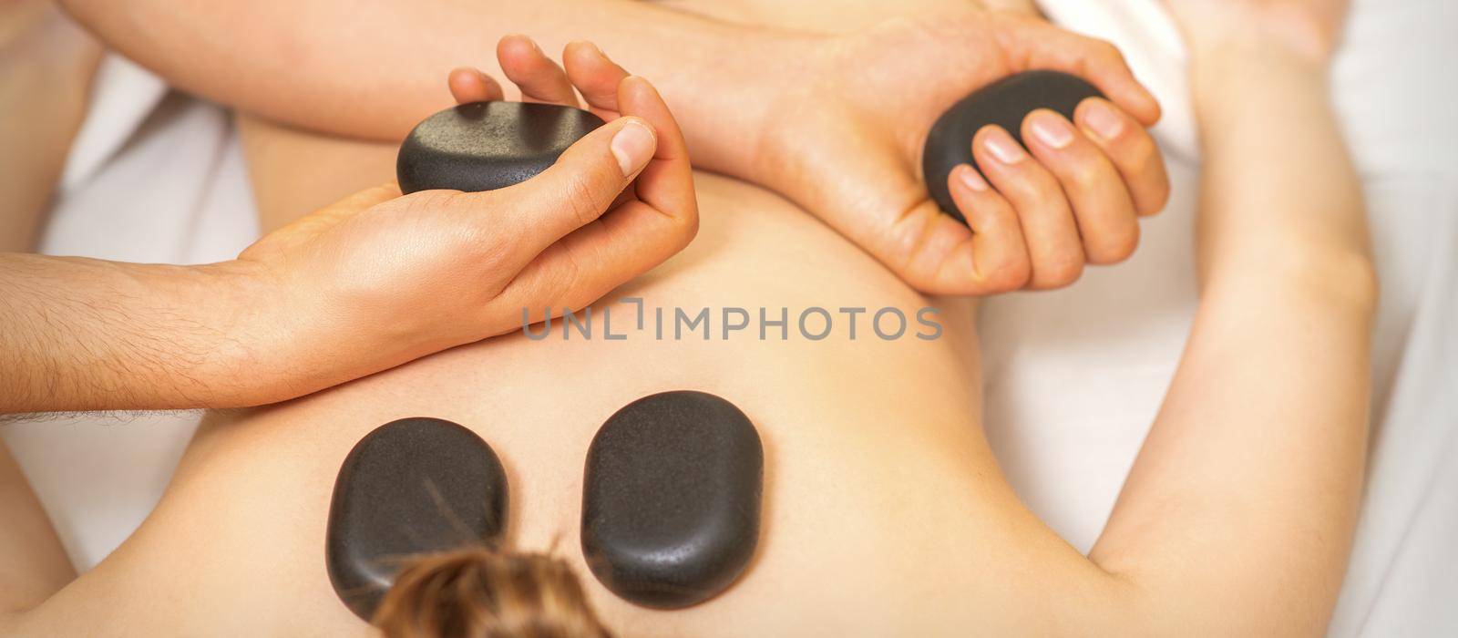 Hot stone massage on the female back with hands of masseur holding black massage stones in spa salon