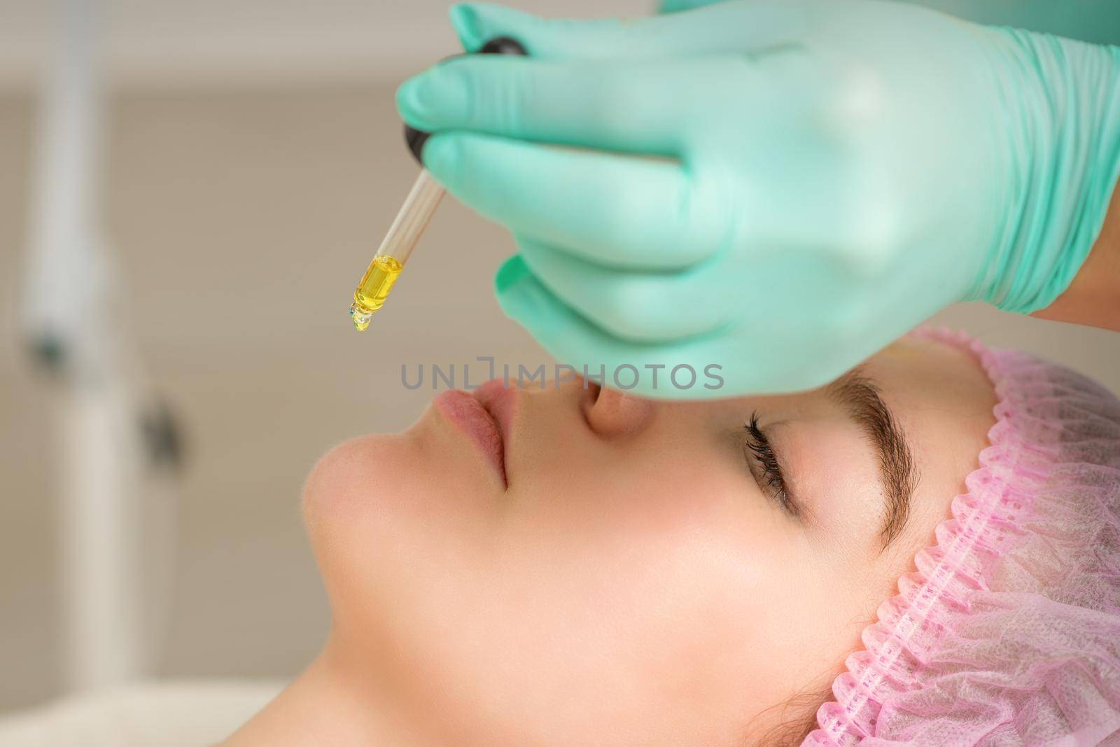 Close up of beautician holding pipette and applying oil serum drop on the face of a young woman in spa salon