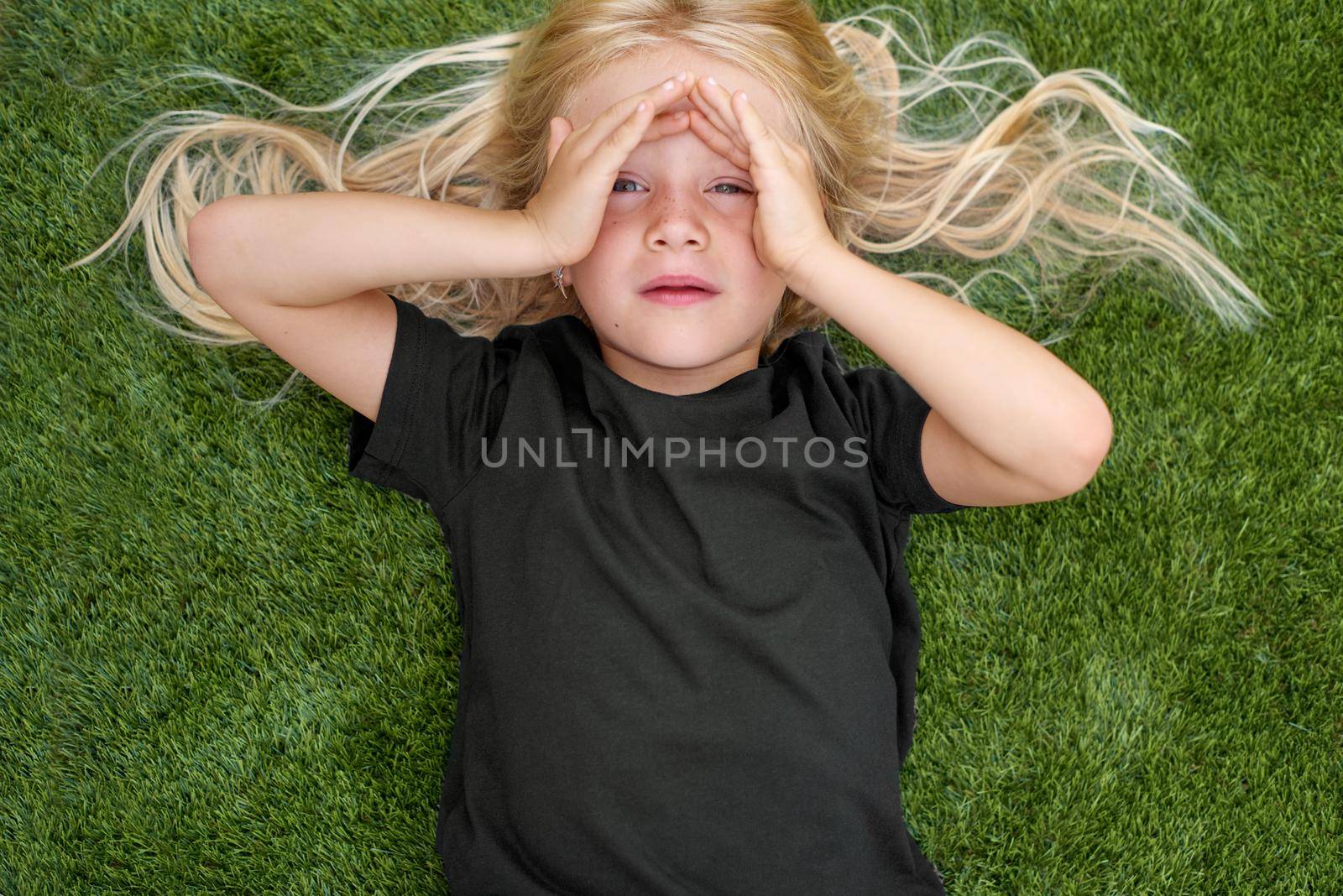 Top view. Mock up. Close up portrait blonde child girl with closed eyes lying, relaxing on green grass. Smiling preschool girl 5 - 6 years old in black t shirt. Lifestyle. Summer vacations. Leisure