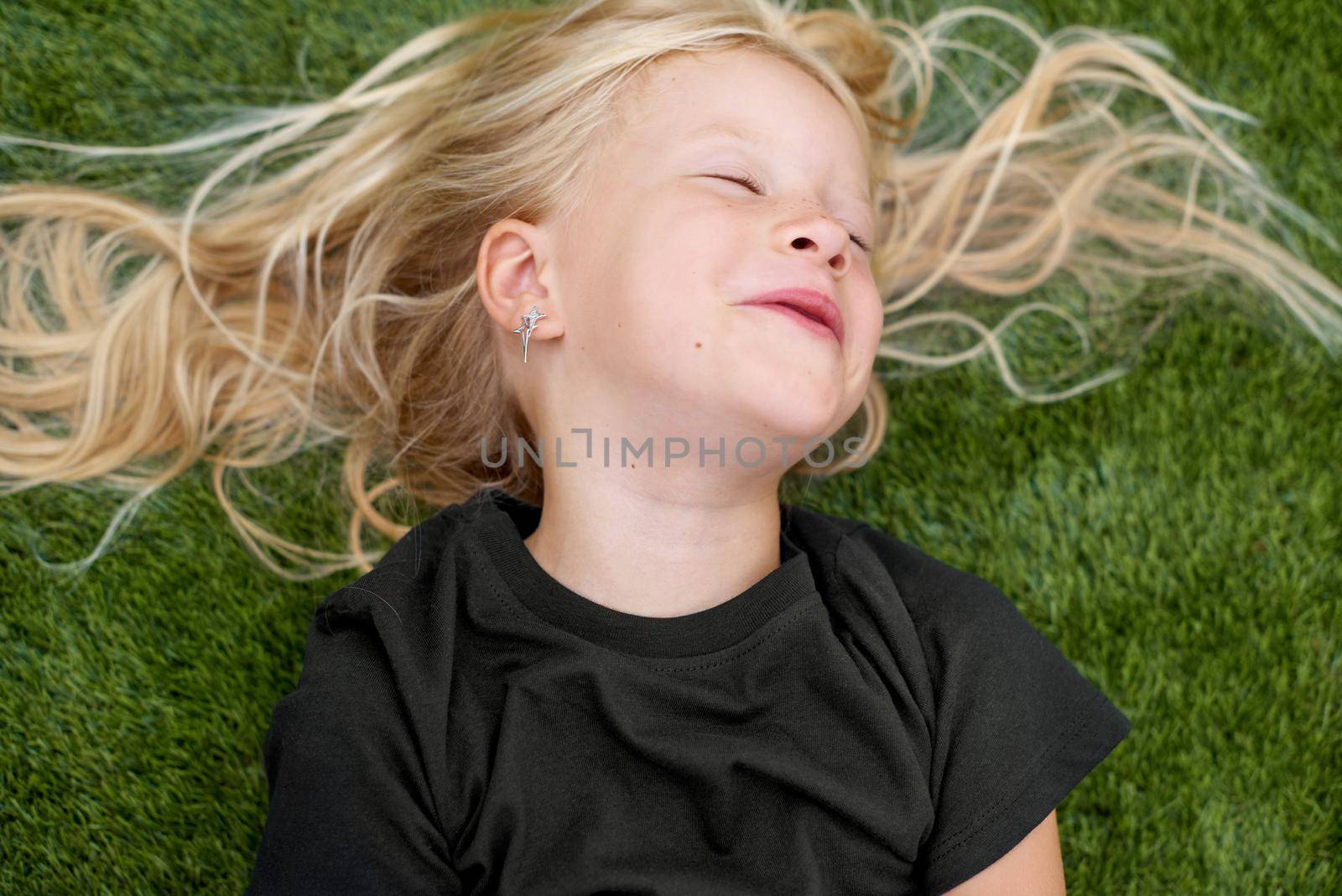 Top view. Mock up. Close up portrait blonde child girl with closed eyes lying, relaxing on green grass. Smiling preschool girl 5 - 6 years old in black t shirt. Lifestyle. Summer vacations. Leisure