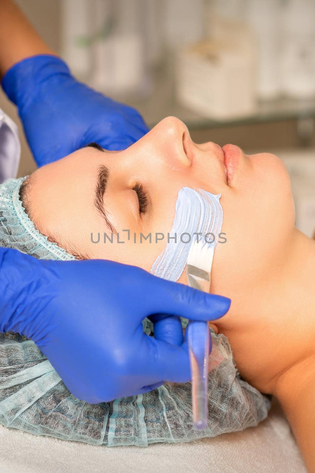 Beautician covering woman facial skin with moisturizing cleansing mask during skin care procedure in a spa beauty salon