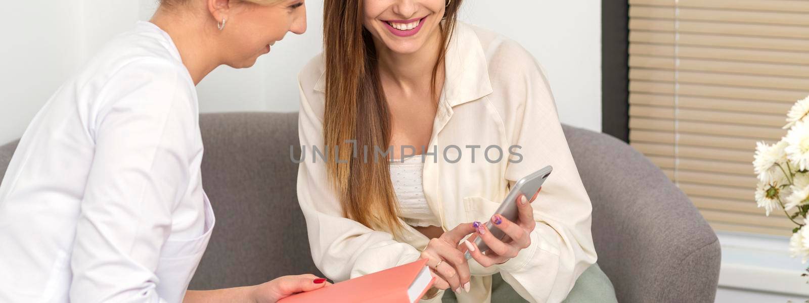 Two young women doctor with pacient smiling and looking at smartphone sitting in hospital office, consultation with a doctor