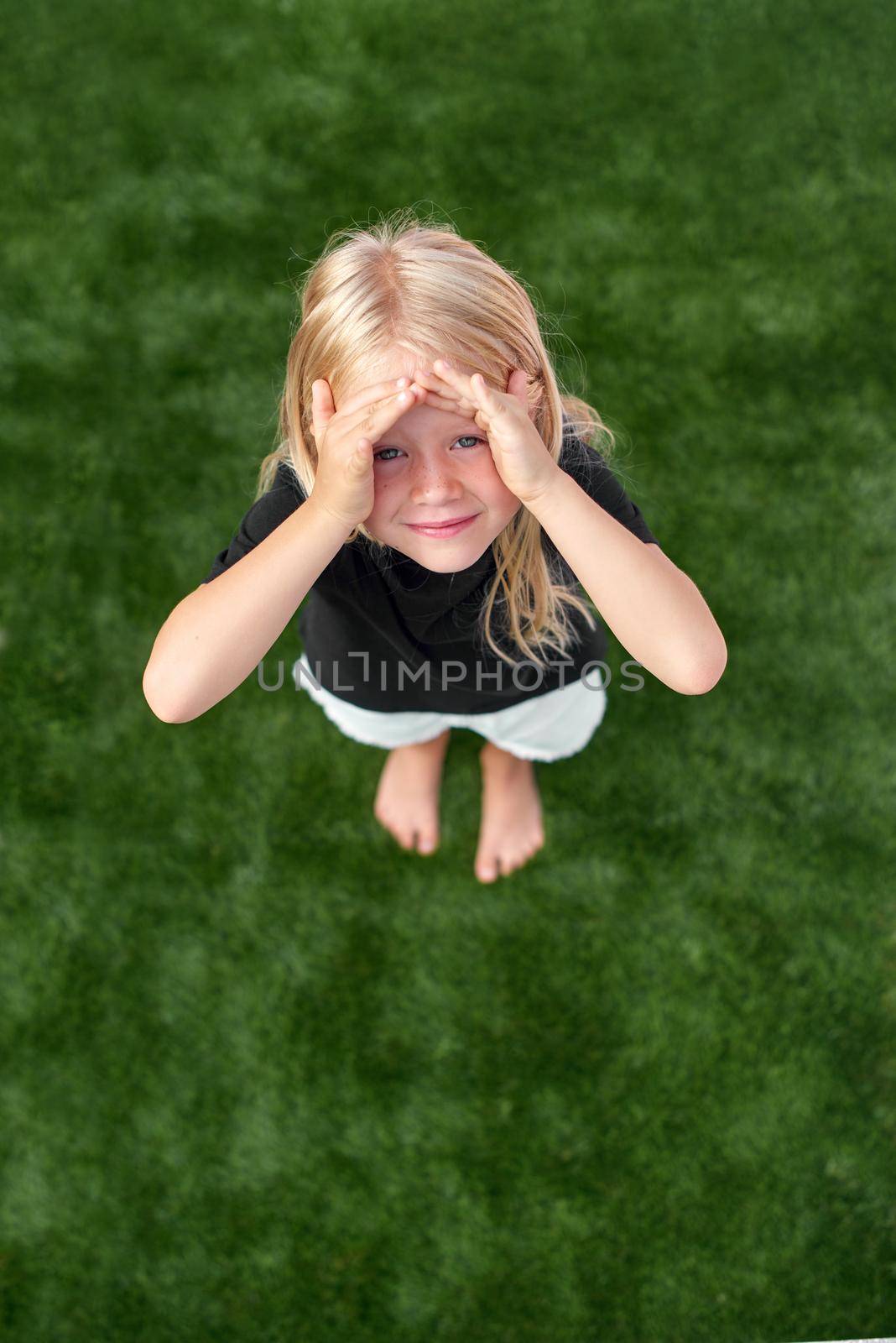 Top view. Mock up. Pretty beautiful blonde child girl standing on green grass. Smiling preschool girl 5 - 6 years old in black t shirt looking at camera. Lifestyle. beauty. Summer vacations. Leisure