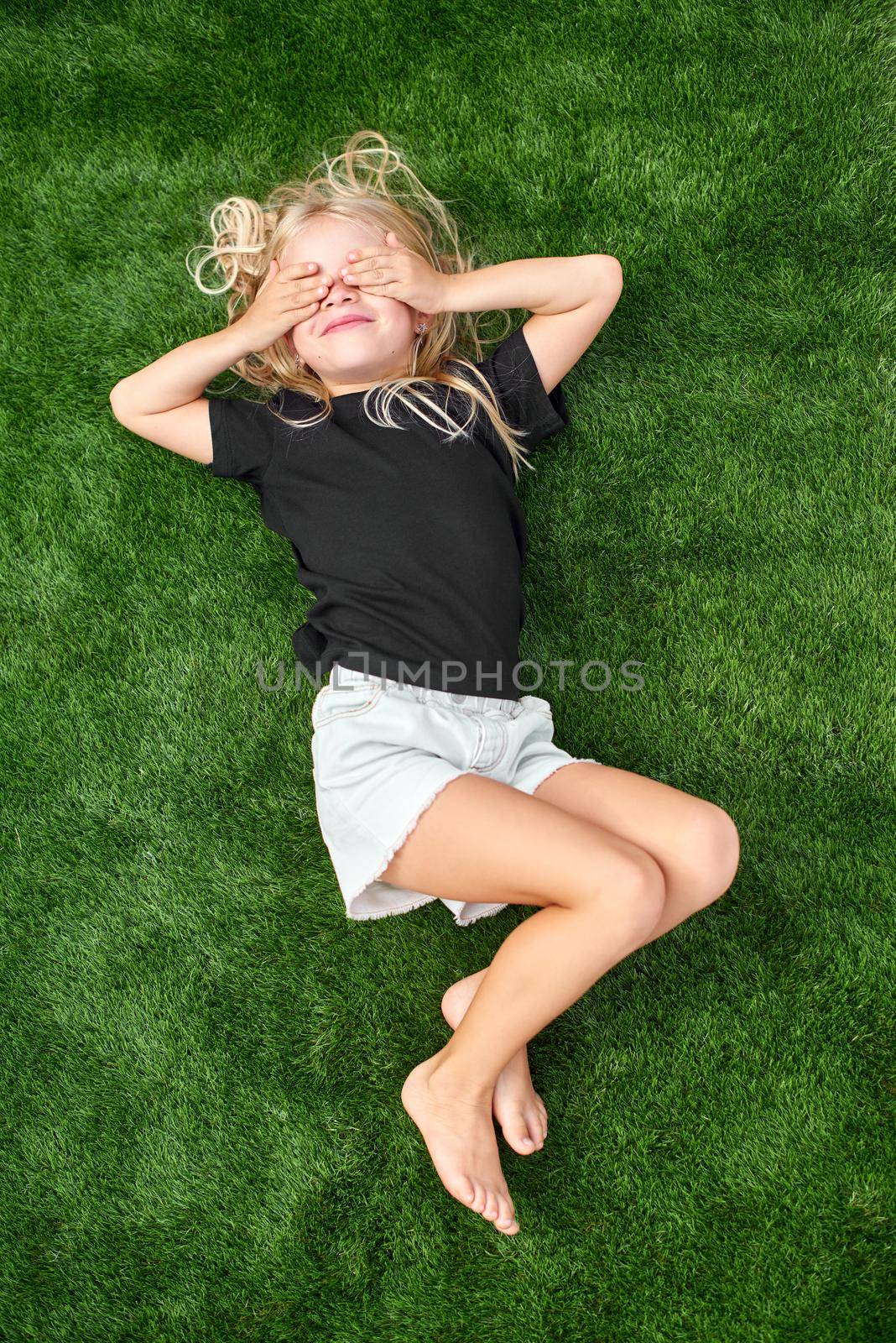 Pretty blonde girl with closed eyes lying and stretching on green grass. Mock up black t shirt. Top view. by OlgaPink